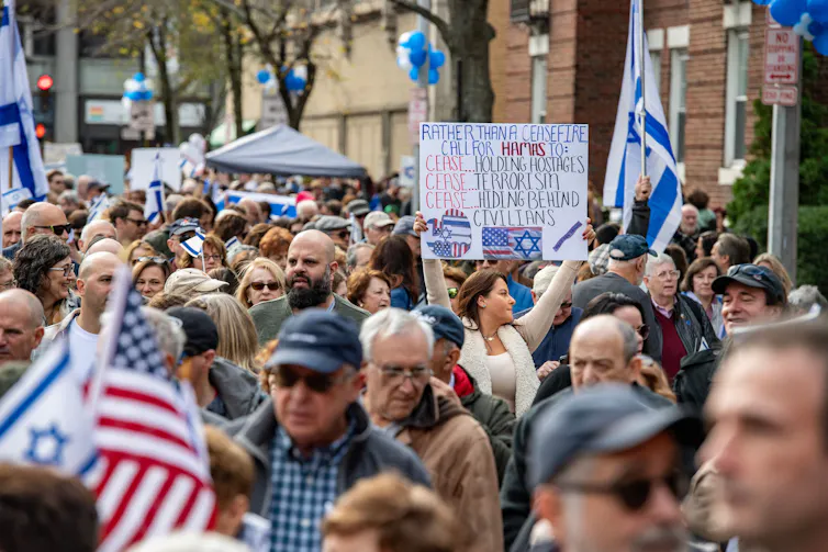 Un gran grupo de personas caminan juntas por la calle, sosteniendo banderas israelíes y estadounidenses y un cartel que dice: 'En lugar de un alto el fuego, pide a Hamás que deje de tomar rehenes, cese el terrorismo, deje de esconderse detrás de los civiles'