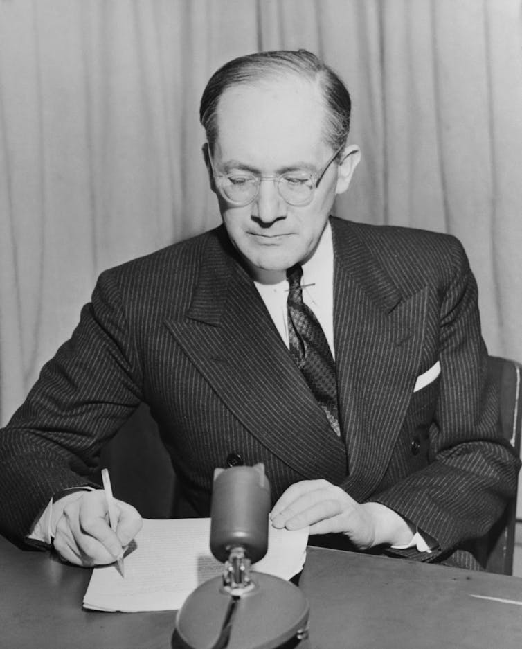 A black and white photo shows a middle aged man wearing a dark suit writing on a piece of paper.