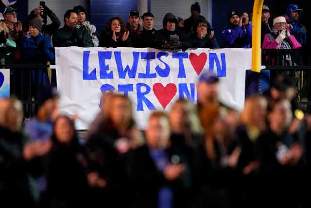 Blurred image of first responders in the foreground standing in front of a sign that reads 'Lewiston Strong.'