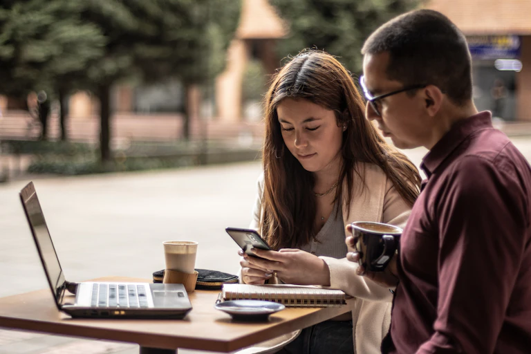 woman and man seated at outdoor table with laptop and looking at phone