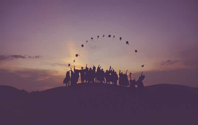 Graduating students toss their caps into the air on a hillside as the sun sets.
