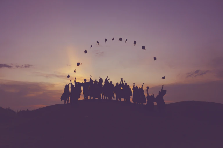 Graduating students toss their caps into the air on a hillside as the sun sets.