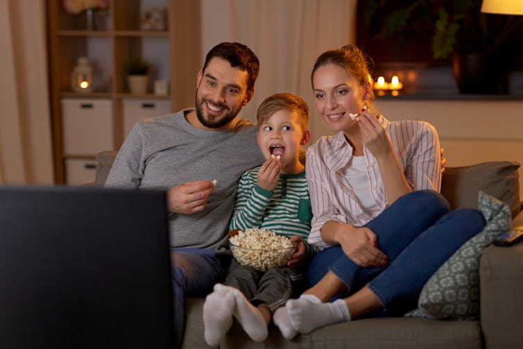 A mother, father and young boy sit on the couch eating popcorn