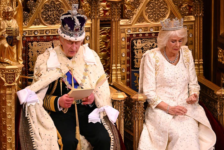 King Charles III and Queen Camilla sitting side by side in their respective thrones while the king reads from a booklet