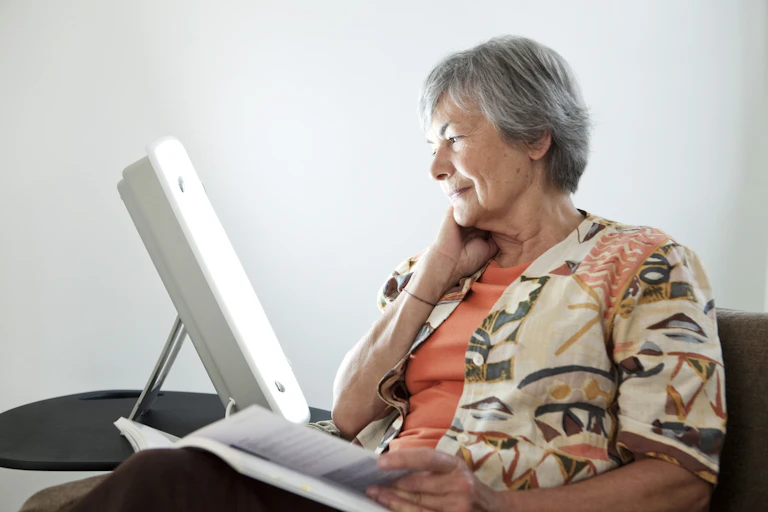 Older woman sits reading by a therapy lamp