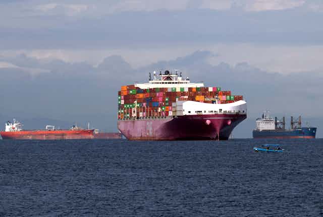 A large cargo ship piled high with goods appears to be in a maritime traffic jam, with four other boats floating nearby.