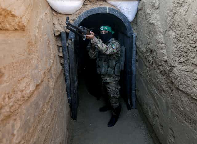 A masked man with a rifle stands in a doorway cut into the ground.