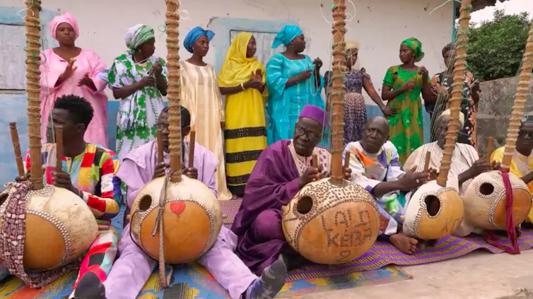 Six hommes vêtus de robes et de coiffes traditionnelles d'Afrique de l'Ouest marchent en groupe en jouant d'instruments à cordes en bois devant une ferme rurale. Derrière eux, une rangée de femmes en tenue traditionnelle applaudissent.