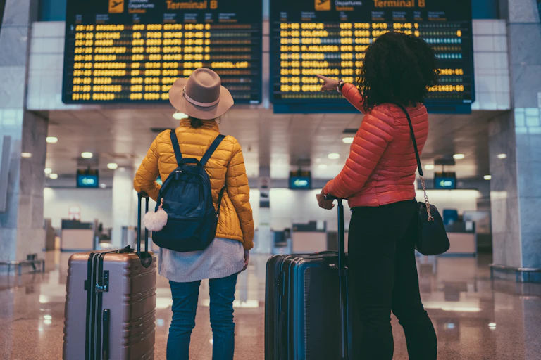 Two women with luggage stand in front of the departures board at an airport.