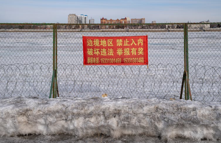 An industrial city is seen in the background behind a fence with Chinese writing on it.