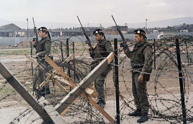 Three soldiers with automatic rifles stand behind a barbed wire fence.