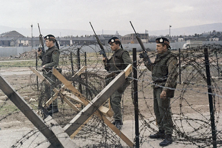 Three soldiers with automatic rifles stand behind a barbed wire fence.