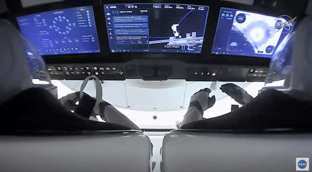 A picture taken in the cockpit of SpaceX's dragon capsule, taken from behind the two pilots, shows white wall out the window. Three screens display text and figures in blue. 