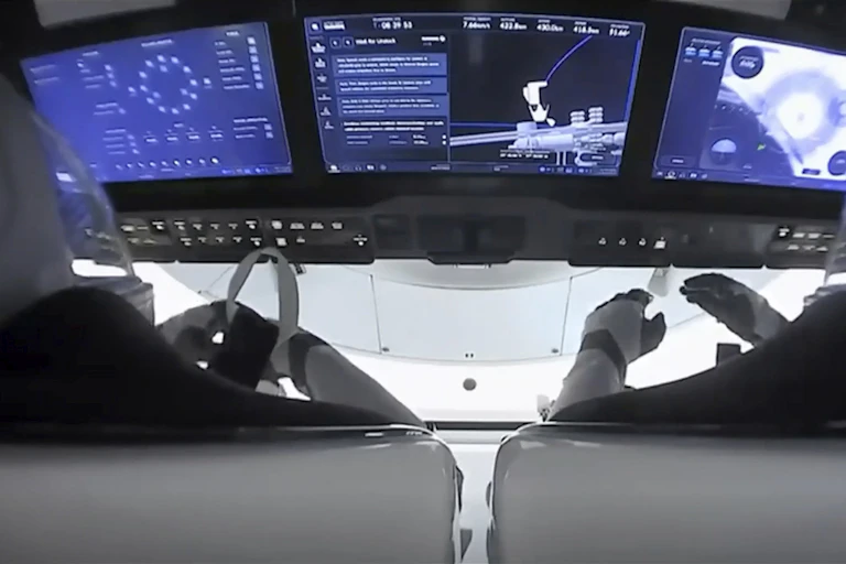 A picture taken in the cockpit of SpaceX's dragon capsule, taken from behind the two pilots, shows white wall out the window. Three screens display text and figures in blue.