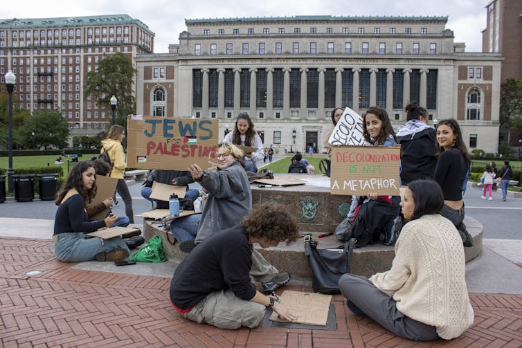People seen sitting at a sculpture, with signs saying decolonization is not a metaphor and Jews for Palestine.