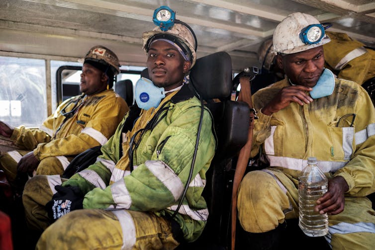 Three men with miners' hats with lights on them and reflective jackets sit in a bus headed for a mine.