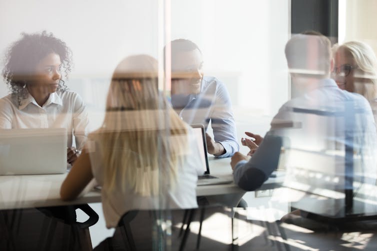 A diverse group of people in business attire sit at a conference table behind a glass door