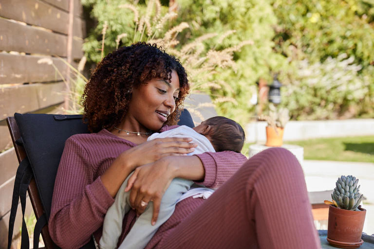Smiling mother lying on a deck chair outside on her patio and looking down at her baby boy in her arms.