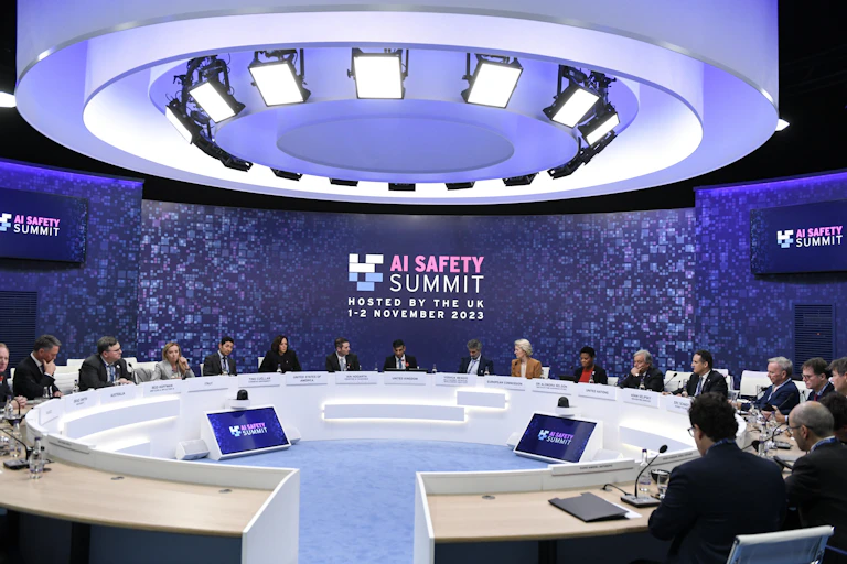 A photo of dignitaries sitting around a conference table in front of a sign that reads 'AI SAFETY SUMMIT, HOSTED BY THE UK 1–2 NOVEMBER 2023"