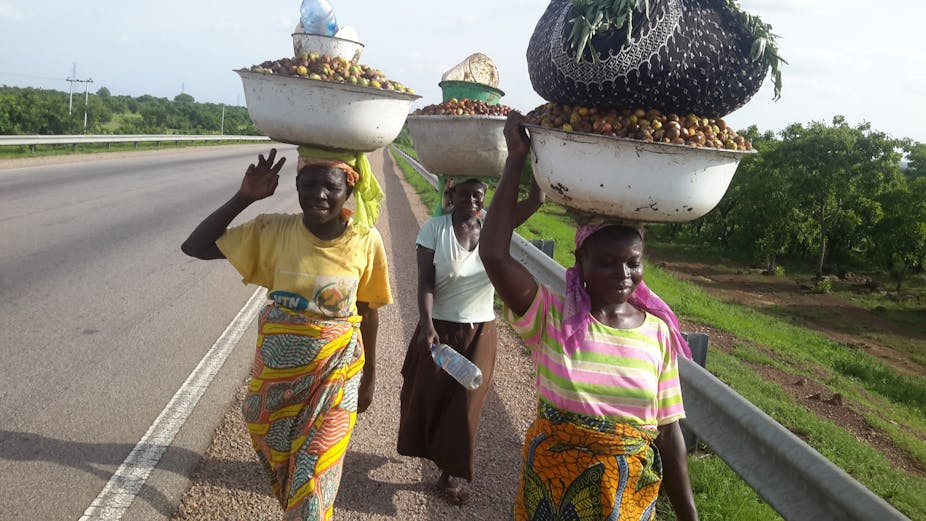 Three women walking along a road with basins of fruit on their heads.