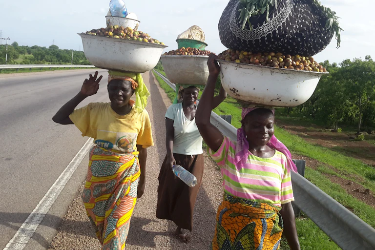 Three women walking along a road with basins of fruit on their heads.