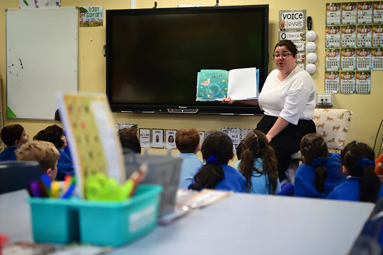 A teacher in a classroom reads to young students who are seated on the floor.