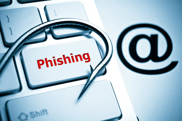 A concept image featuring a keyboard on which the word "phishing" is written with a fishing hook laid atop it, alongside an "at" symbol