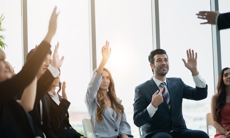 Group of people in suits with raised hands, man in suit pointing to himself.