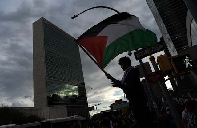 A person waves a Palestinian flag in front of a tall building.