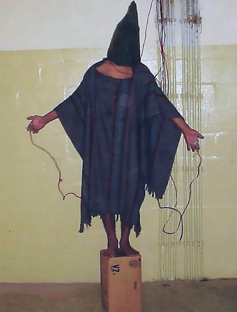 A man standing on a box wearing a hood and with his hands held out, apparently attached to power cables.
