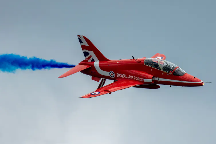 Small red aircraft with Union Flag on its tail and blue smoke from its exhaust.