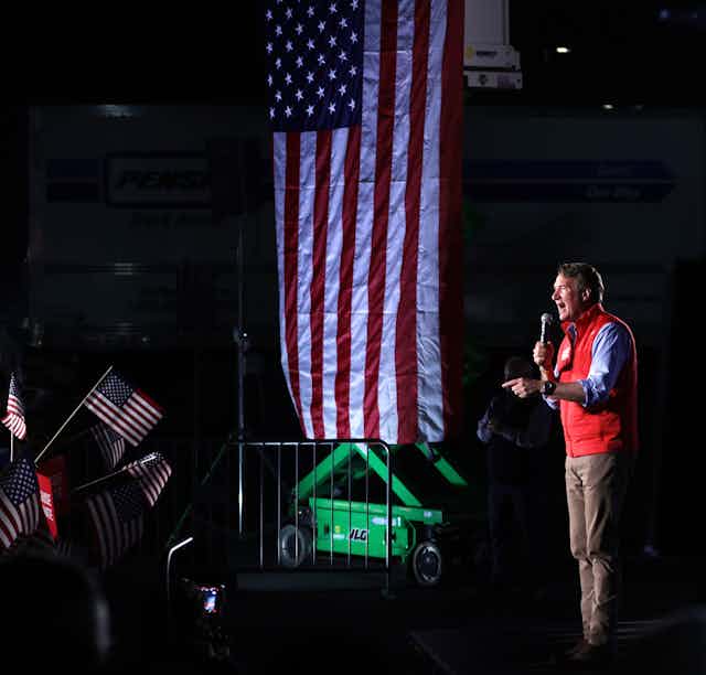 A man wearing a red vest is holding a microphone as he stands near an American flag and a crowd of people. 