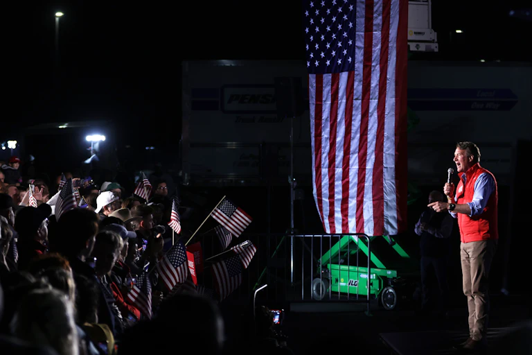 A man wearing a red vest is holding a microphone as he stands near an American flag and a crowd of people.