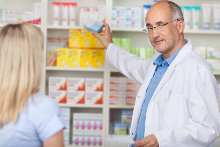 Une femme de dos est devant le comptoir d'une pharmacie, face à un pharmacien qui lui tend un médicament.