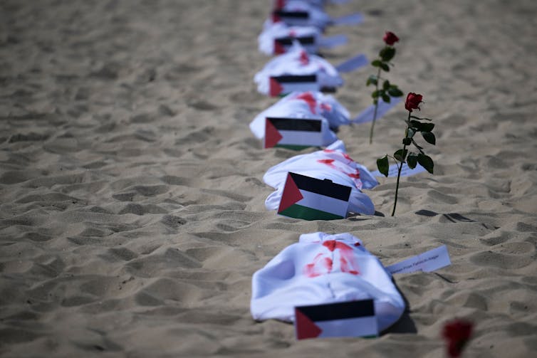 Mantas blancas con pintura roja se encuentran junto a banderas palestinas en una playa.
