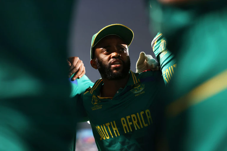 A man in a green and gold jersey and peak cap is seen through bodies huddled together, talking earnestly.