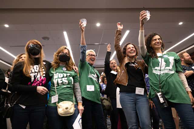 Six women raise their glasses and hands and pose together as they wear green and black shirts that say 'vote Yes 1.'
