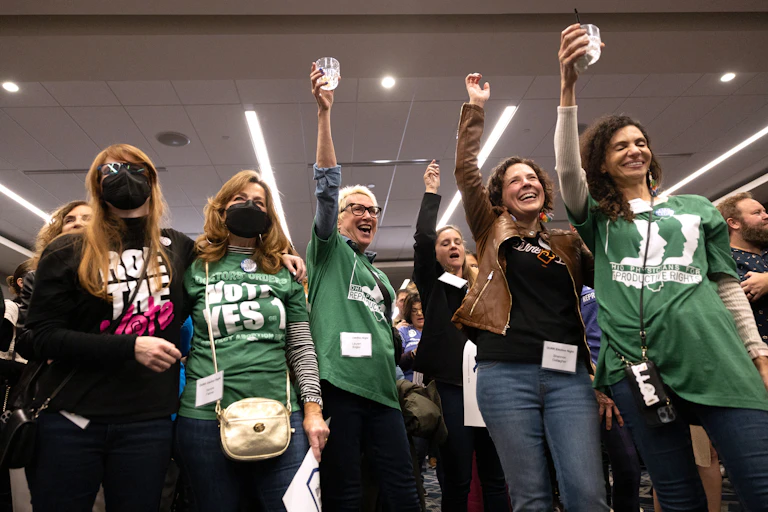 Six women raise their glasses and hands and pose together as they wear green and black shirts that say 'vote Yes 1.'