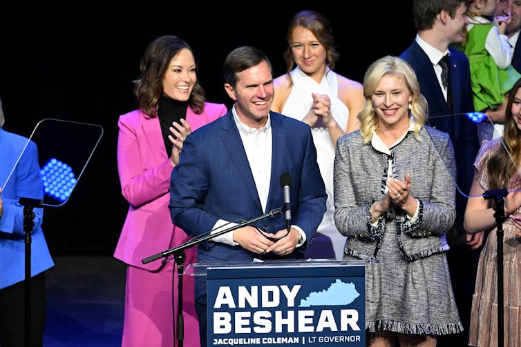 Andy Beshear stands in a dark blue suit at a podium that has his name on it, surrounded by three women on a stage.