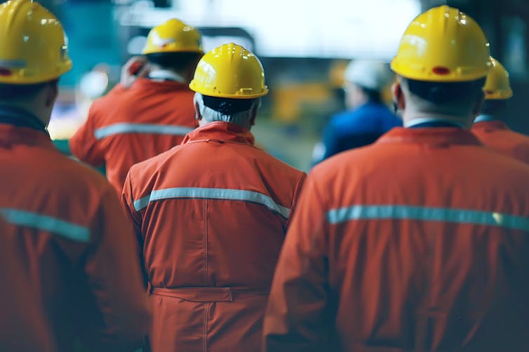 Workers in orange overalls and yellow hard hats stand with their backs to the camera.