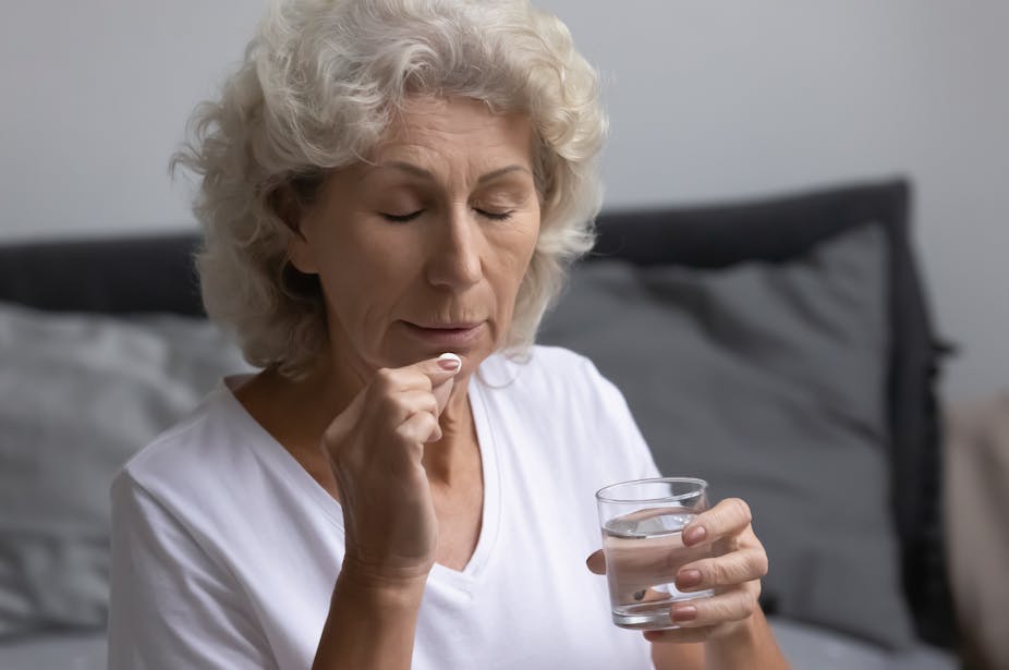 An older woman holds a glass of water while placing a pill in her mouth.