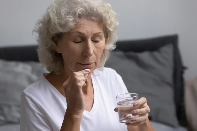 An older woman holds a glass of water while placing a pill in her mouth.
