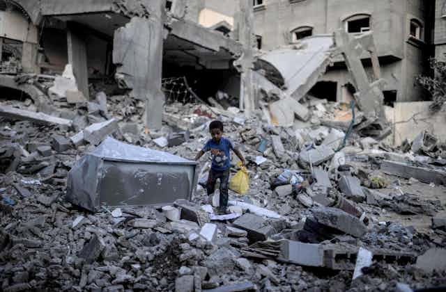 A young boy holding a yellow bag walks through rubble.