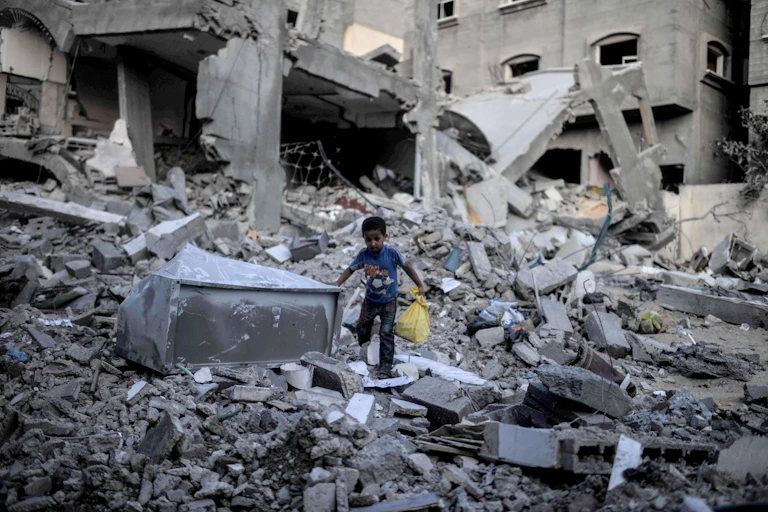 A young boy holding a yellow bag walks through rubble.