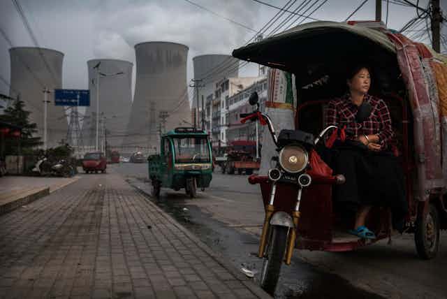 A Chinese vendor waits for customers as she sells vegetables at a local market outside a state owned coal fired power plant in Huainan, China. The power plants large towers are in the background.