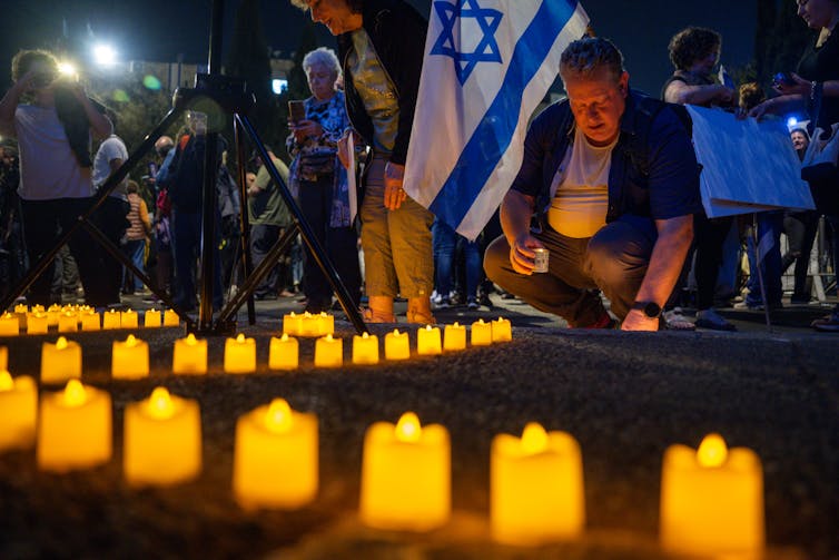 A man kneels by a row of candles. He is behind a blue and white Israeli flag.