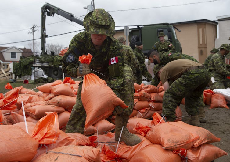 Soldiers in camouflage clothing handle orange sandbags.