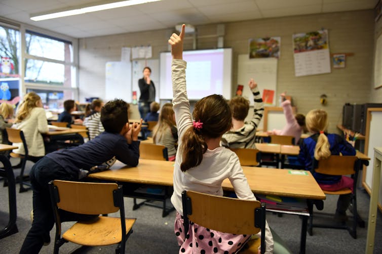 A classroom of children raise their hand, looking at the teacher.