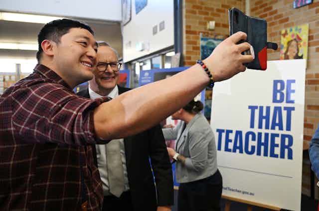 A man takes a selfie with Anthony Albanese in front of a sign that says 'Be That Teacher'. 