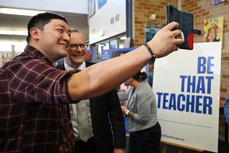 A man takes a selfie with Anthony Albanese in front of a sign that says 'Be That Teacher'.